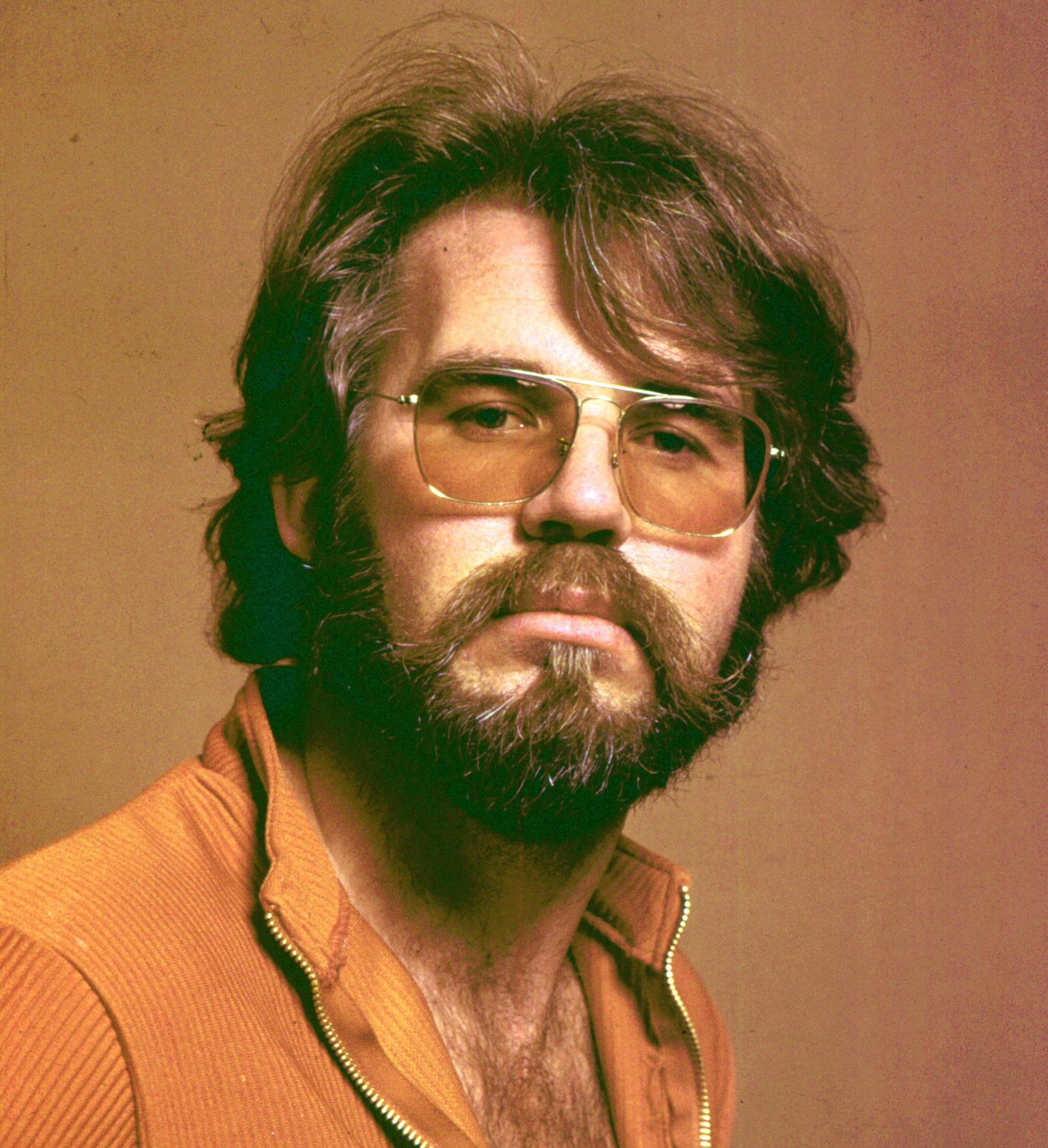 Kenny Rogers looks directly into the camera in a vintage 1960s studio portrait, sporting his signature shaggy brown hair, full beard, and aviator glasses with tinted yellow lenses. The singer-songwriter wears a casual ribbed orange zip-up top slightly unzipped at the chest against a neutral, warm-toned backdrop. | Source: Getty Images