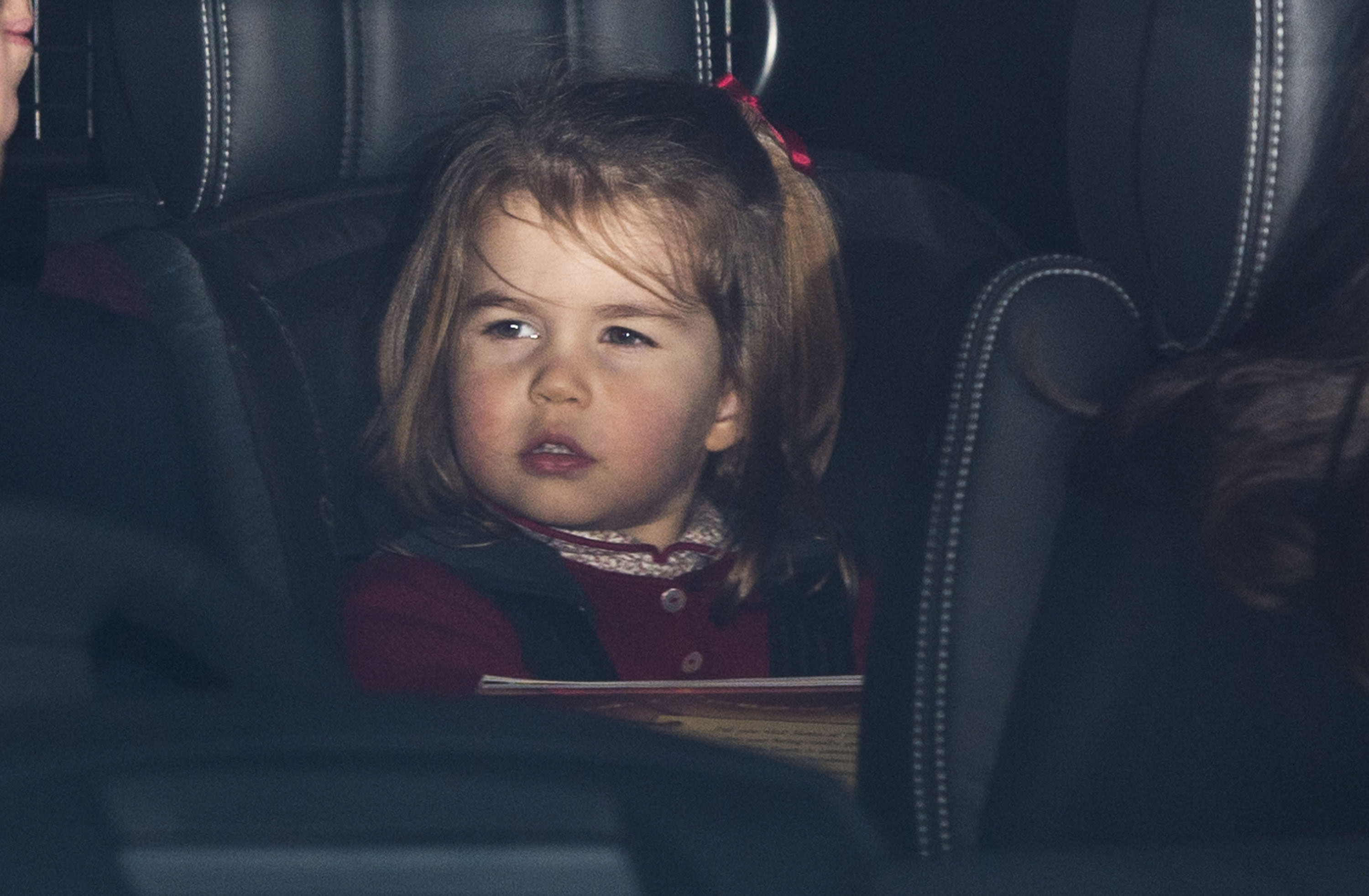 Princess Charlotte of Cambridge peers out from the back seat of a car arriving for the annual Christmas lunch for the extended Royal Family at Buckingham Palace on December 20, 2017, in London, England. The young royal wears a festive burgundy cardigan over a patterned collared top, with her hair styled with a matching red ribbon bow. | Source: Getty Images