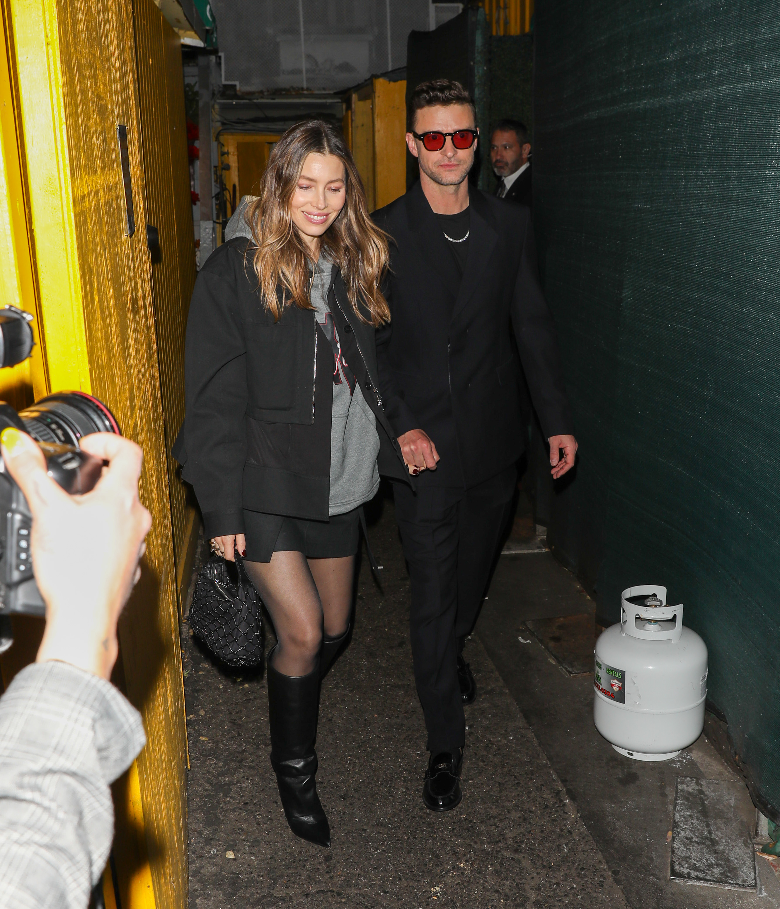 Jessica Biel and Justin Timberlake hold hands while walking through a dimly lit passage. Biel wears a black jacket over a gray top, a short black skirt, sheer tights, and knee-high boots, while Timberlake opts for an all-black outfit with red-tinted sunglasses.