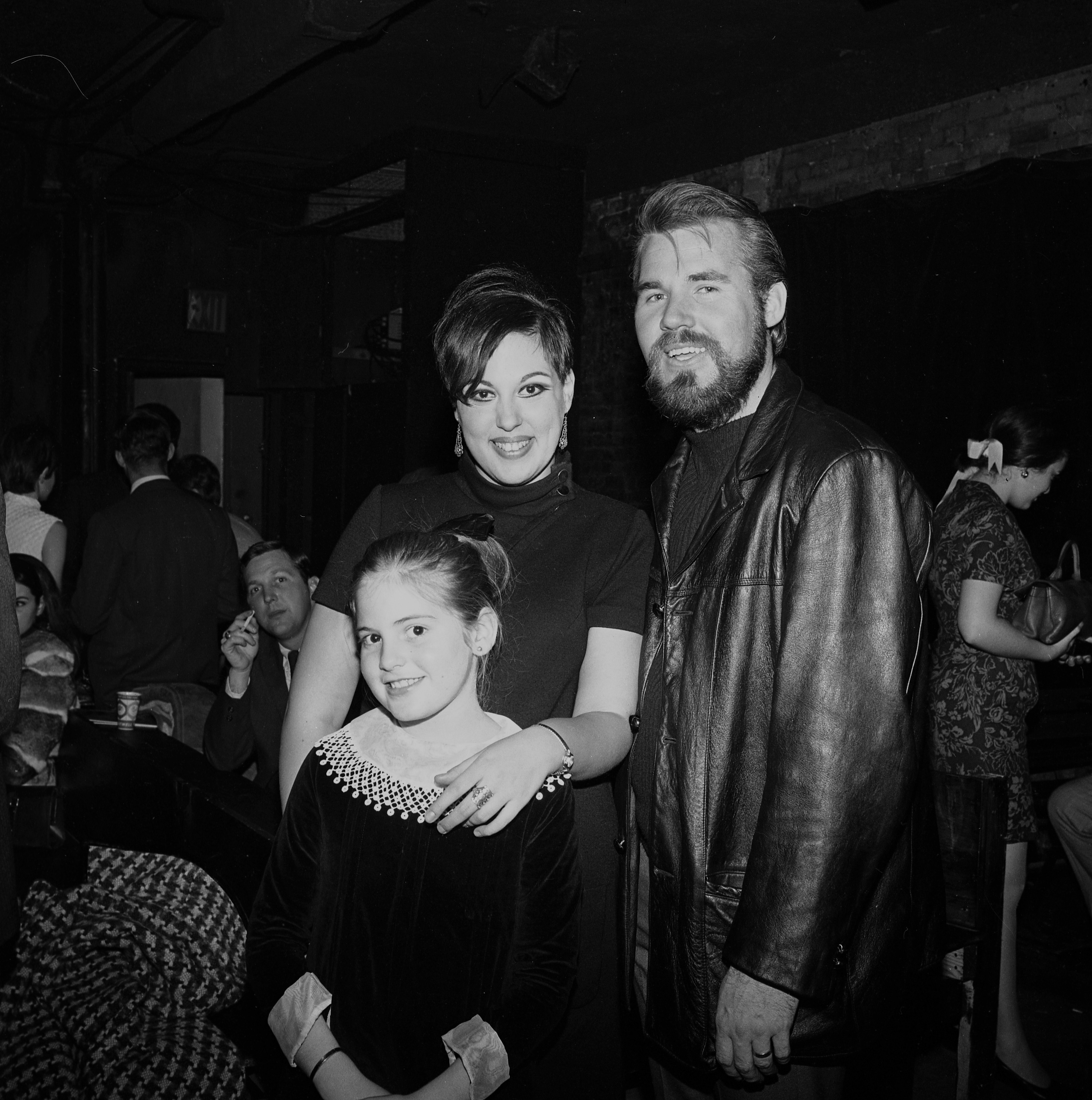 Kenny Rogers, then bass player and singer for The First Edition, poses alongside an unidentified woman and young girl at The Bitter End nightclub on November 8, 1967, in New York City. Sporting a dark leather coat over a turtleneck, Kenny smiles as the woman rests a hand on the smiling young girl in a velvet dress with white lace trim. | Source: Getty Images