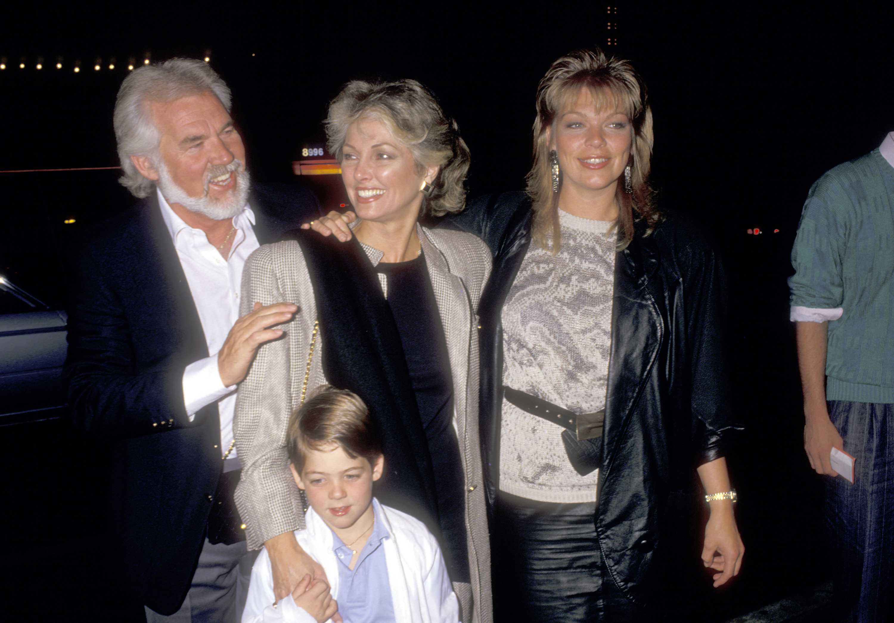 Kenny Rogers enjoys a family outing with his wife Marianne Gordon, young son Christopher Cody Rogers, and daughter Carole Rogers at the David Copperfield Magic Show on May 15, 1987, in Hollywood, California. Kenny laughs warmly in a dark blazer and open white collared shirt as Marianne stands beside him in a tweed coat over a black top, holding hands with little Christopher, while Carole beams on the right in a stylish black leather jacket and patterned knit sweater. | Source: Getty Images