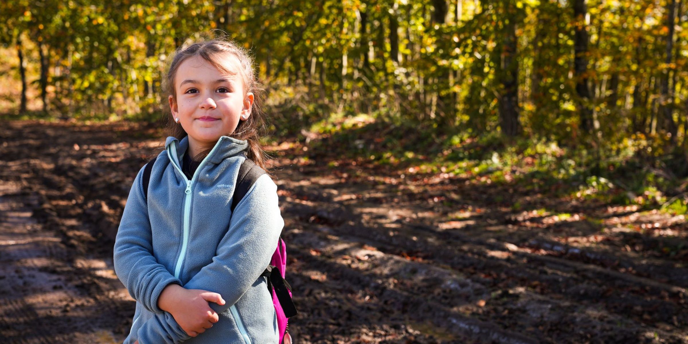Young girl wearing a blue jacket and backpack standing on a forest trail | Source: Pexels