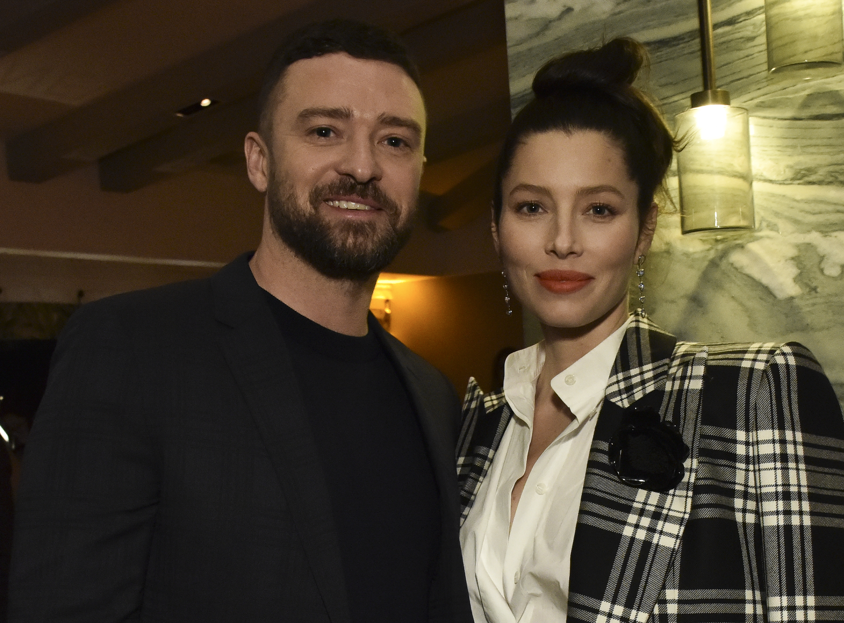 Justin Timberlake and Jessica Biel pose together indoors, smiling at the camera. Timberlake wears a dark blazer over a black shirt, while Biel pairs a black-and-white plaid blazer with a white blouse and a black floral accessory.