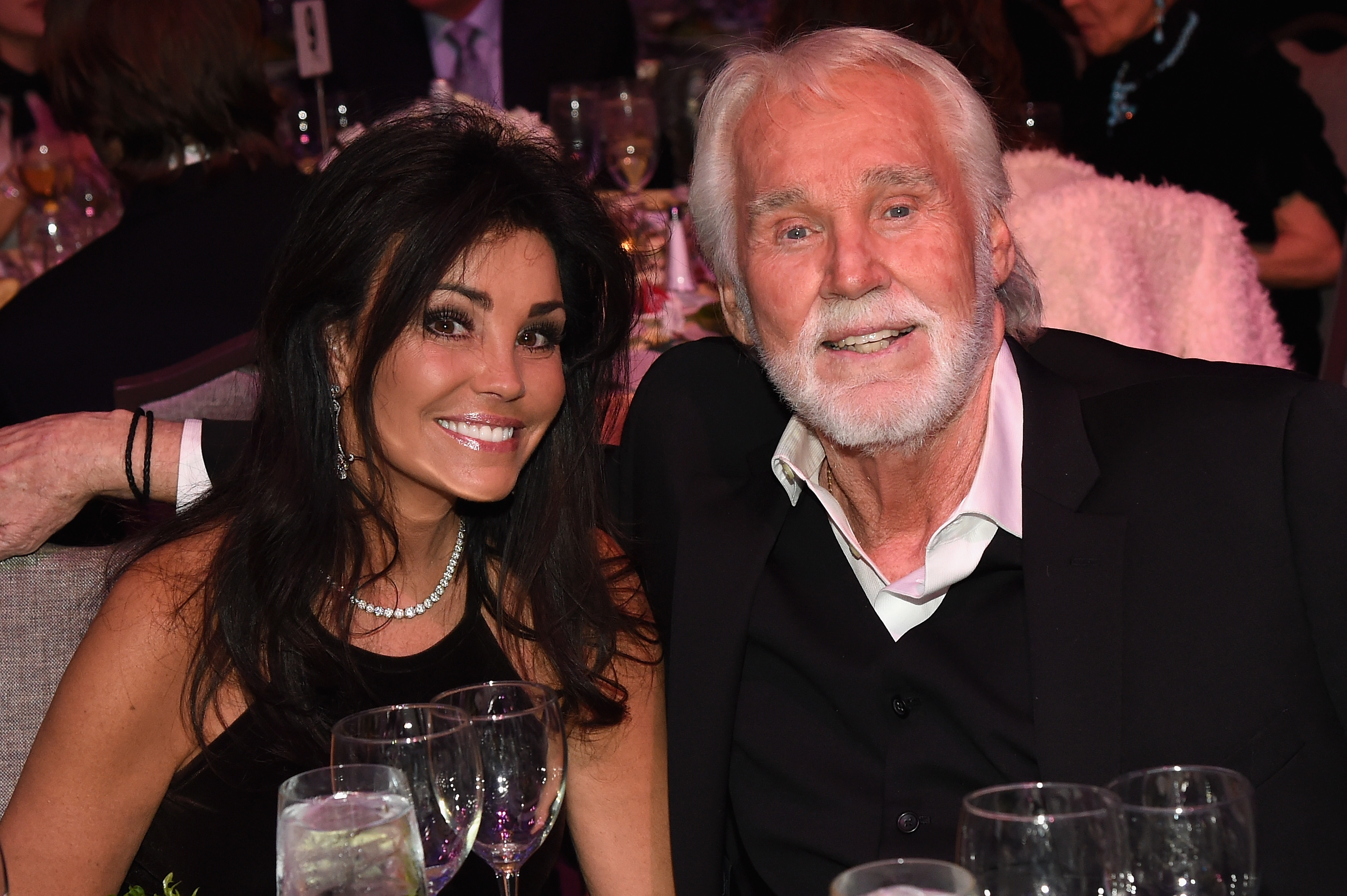 Kenny Rogers and his fifth wife, Wanda Miller, smile together at their table during the T.J. Martell Foundation 8th Annual Nashville Honors Gala at the Omni Nashville Hotel on February 29, 2016. Wanda wears an elegant black sleeveless dress accessorized with a diamond necklace and drop earrings, while Kenny puts his arm around her, dressed in a black suit with a white collared shirt. | Source: Getty Images