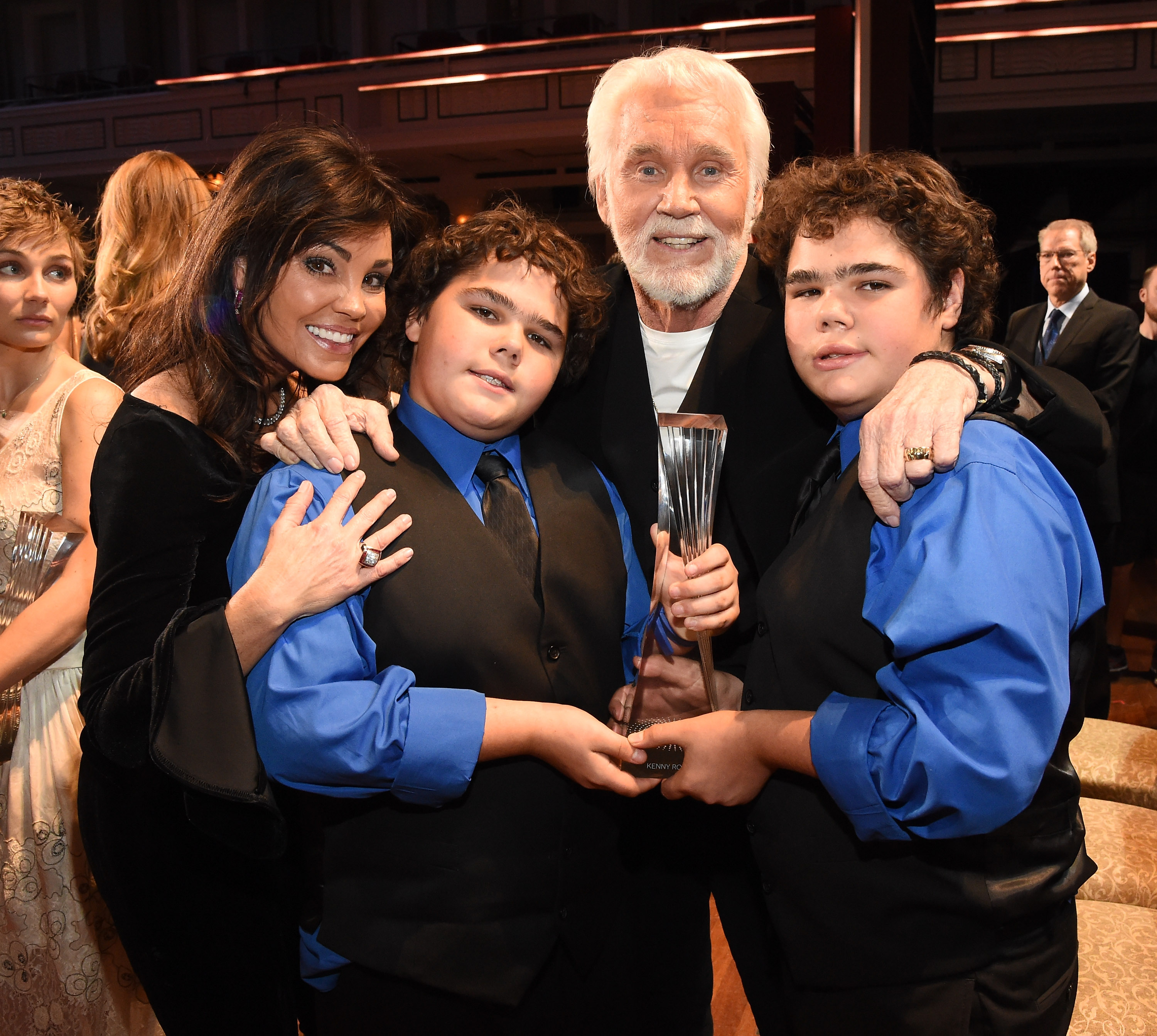 Honoree Kenny Rogers celebrates with his wife Wanda Miller and their twin sons, Justin and Jordan, at the 2015 "CMT Artists of the Year" ceremony at the Schermerhorn Symphony Center on December 2, 2015, in Nashville, Tennessee. The boys, dressed in matching blue shirts, black vests, and ties, proudly help hold their father's crystal trophy as the family embraces and smiles for the camera.
