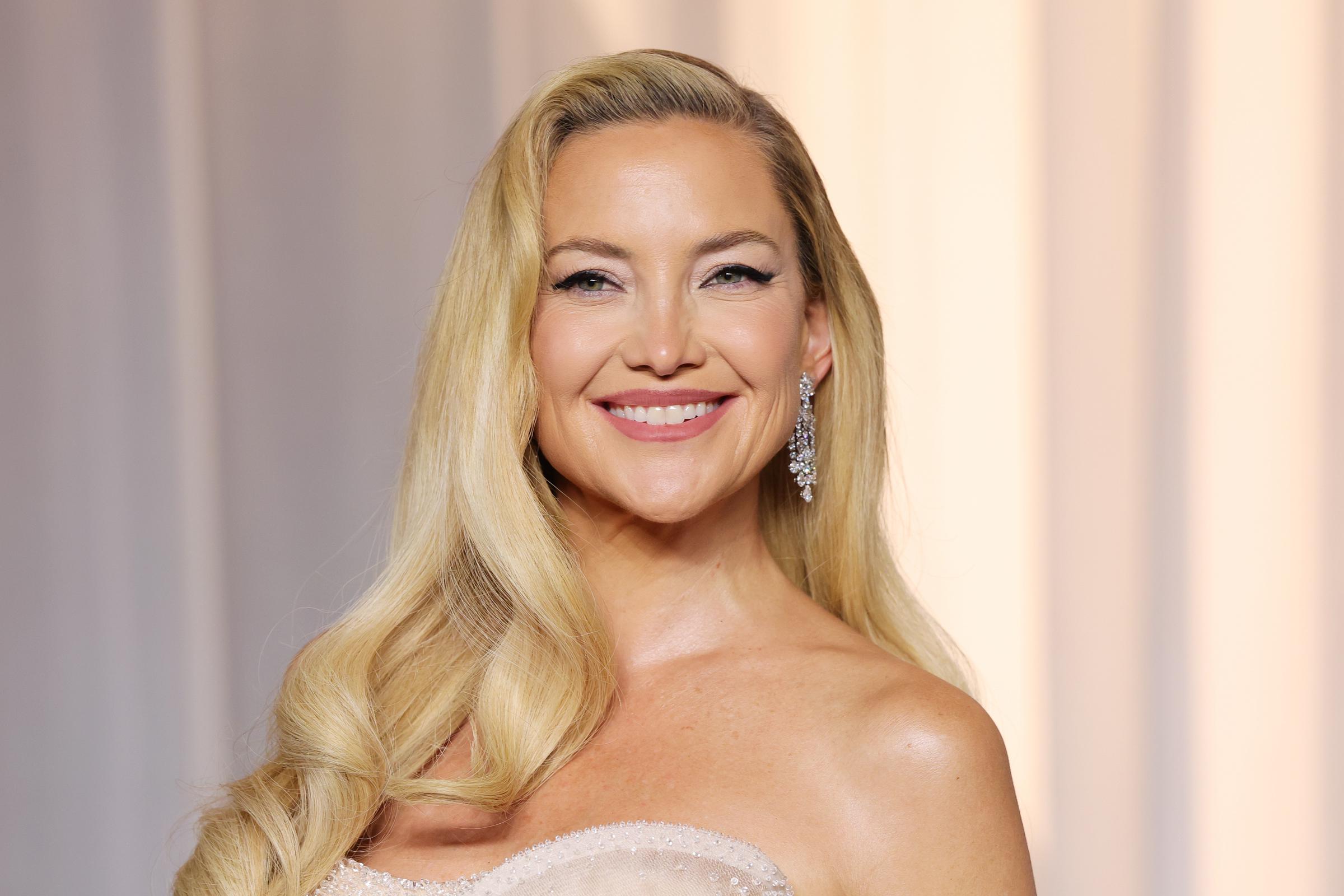 Kate Hudson smiles for a close-up portrait, pairing her side-swept blonde waves with sparkling drop earrings, winged eyeliner, and soft pink lipstick.