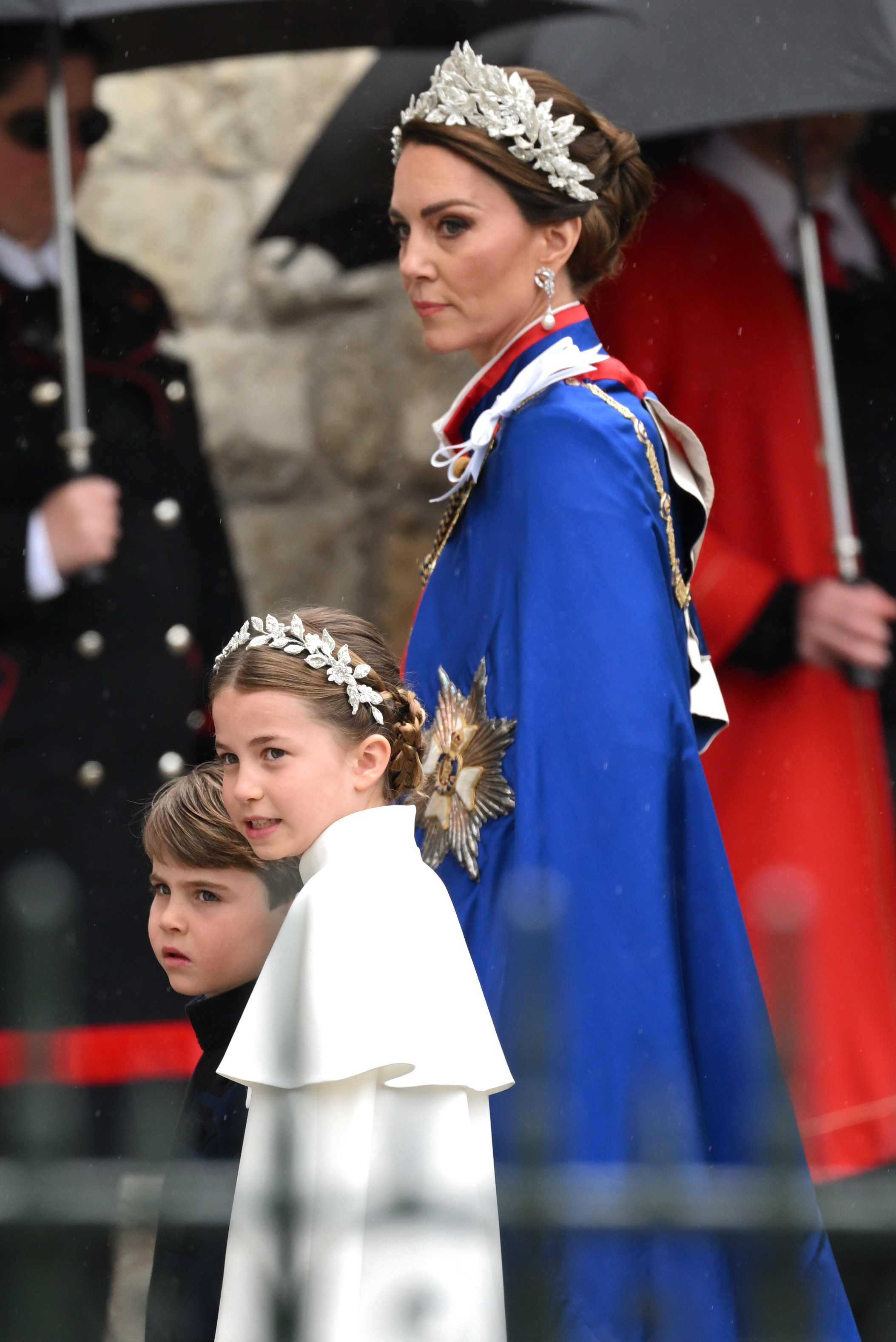 Princess Charlotte arrives alongside Prince Louis and Catherine, Princess of Wales at Westminster Abbey for the Coronation of King Charles III and Queen Camilla on May 6, 2023, in London, England. Charlotte mirrors her mother in an ivory Alexander McQueen dress with a capelet and an intricate silver bullion and crystal flower headpiece, walking alongside Prince Louis in a navy Dege & Skinner tunic while Catherine stands regally in the Royal Victorian Order mantle and a matching floral headpiece. | Source: Getty Images