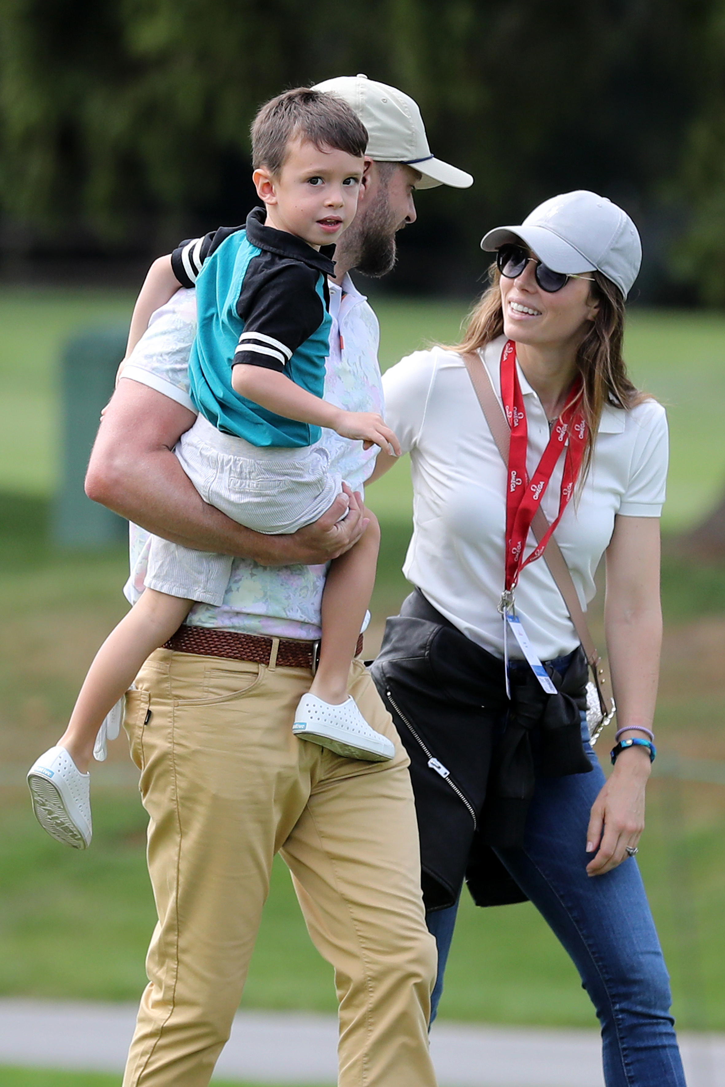 Justin Timberlake carries his young son while walking outdoors with Jessica Biel. The singer wears a light-colored cap and pastel-patterned polo shirt, while Biel sports a white cap, sunglasses, a white polo shirt, and jeans.
