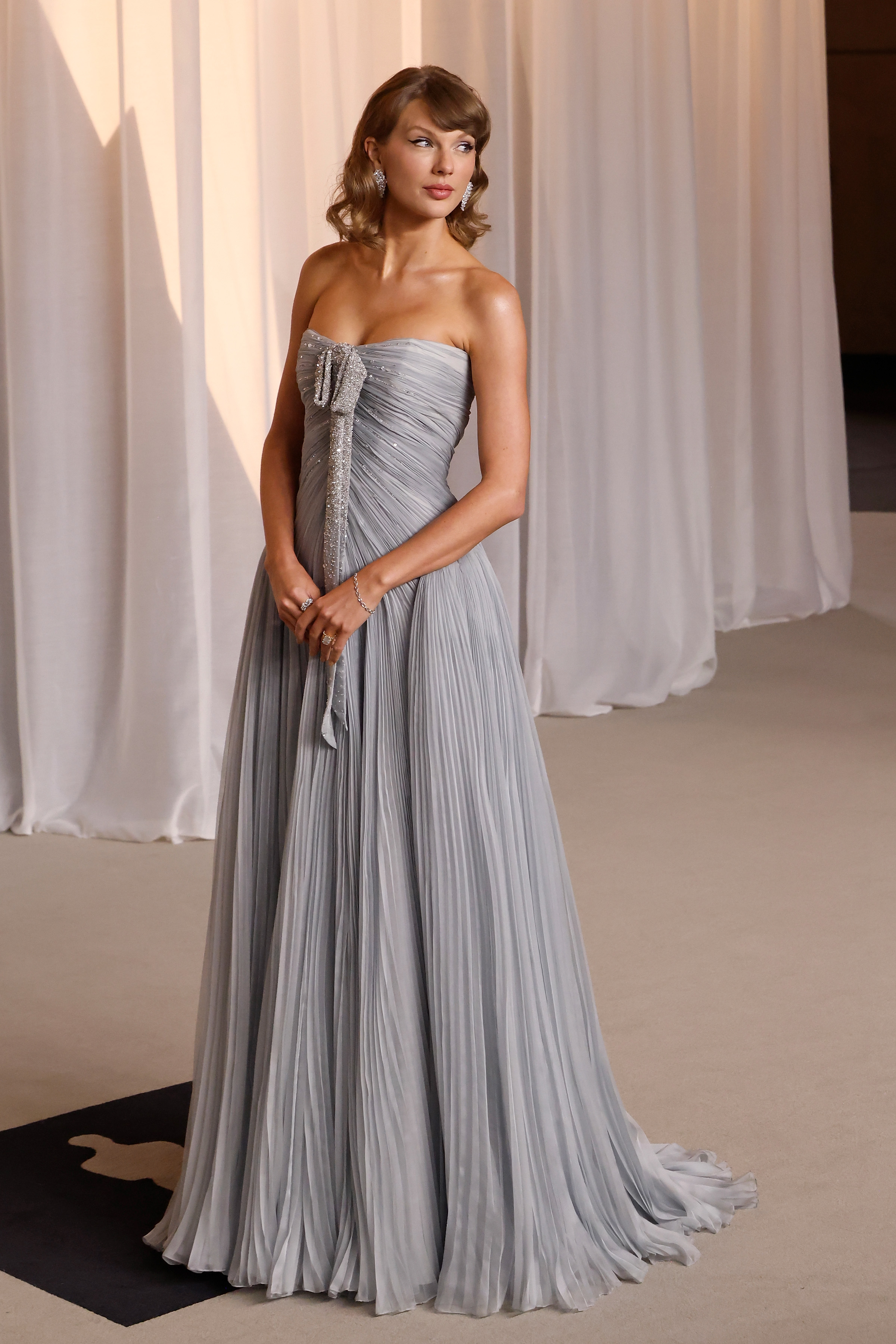 Swift poses at an angle in the flowing strapless gown, showing its sweeping silhouette and train while her curled, shoulder-length hair frames her face.