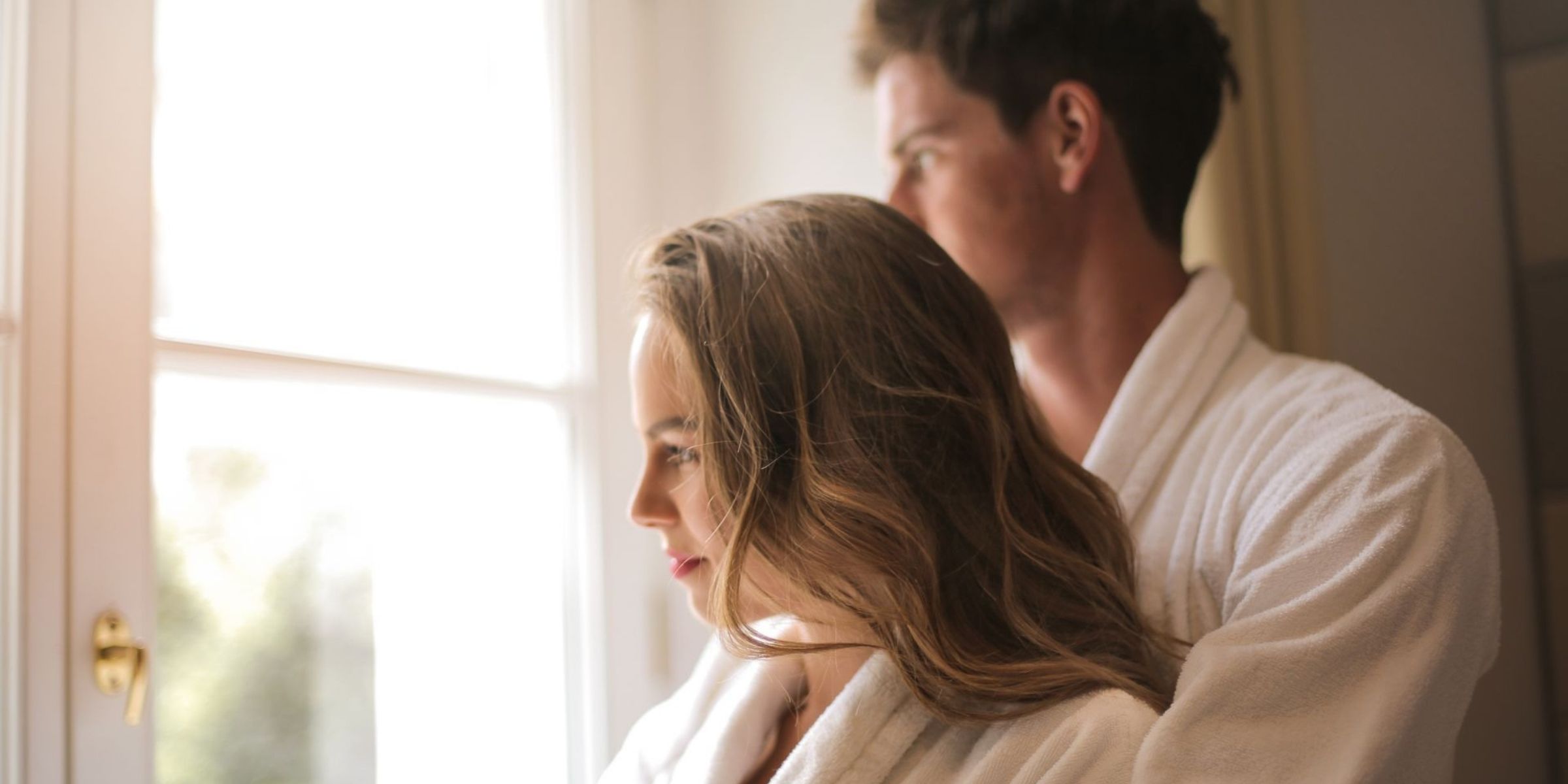 Couple wearing white bathrobes standing together by a sunlit window | Source: Pexels