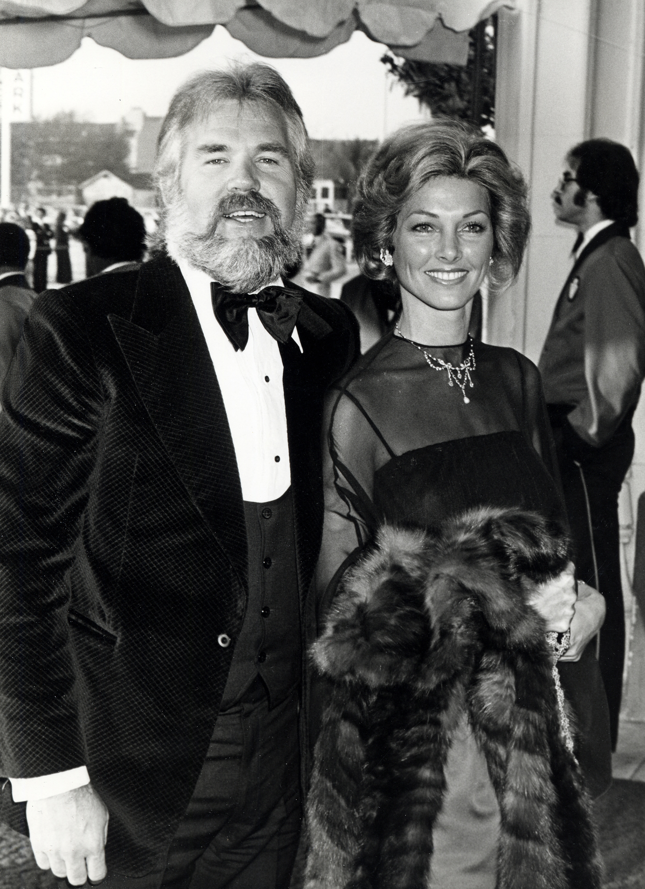 Kenny Rogers and his fourth wife, actress Marianne Gordon, arrive in style at the 20th Annual Grammy Awards at the Shrine Auditorium on February 23, 1978, in Los Angeles. Kenny smiles in an elegant black velvet tuxedo with a matching vest, bow tie, and white dress shirt, while Marianne dazzles beside him in a sheer-sleeved black evening gown accented with a sparkling statement necklace and a luxurious fur wrap draped over her arm. | Source: Getty Images
